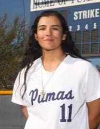 A woman in white baseball uniform standing next to a field.